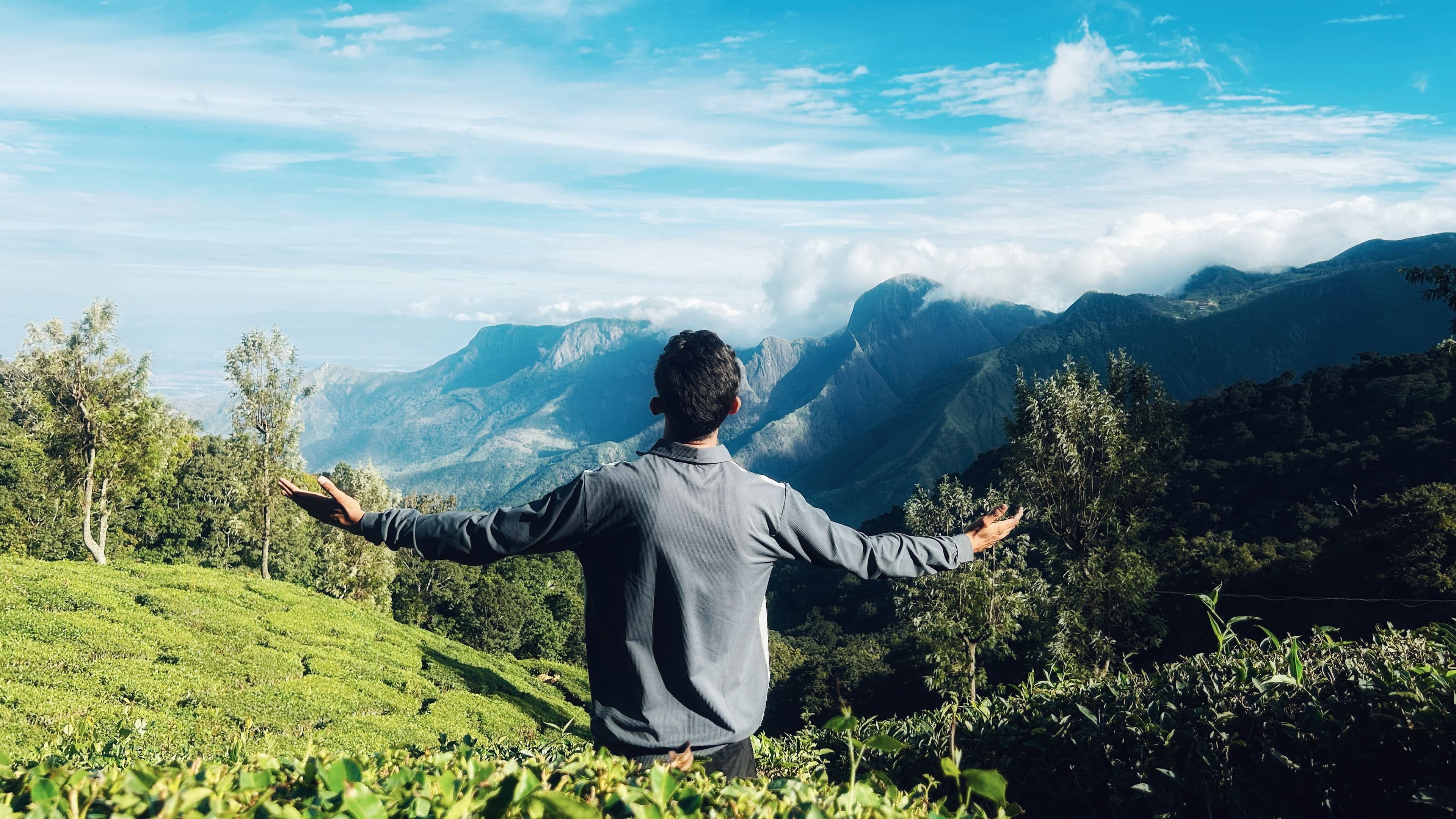 A man with open arms enjoying the breathtaking view of Munnar's lush tea plantations and majestic mountains.